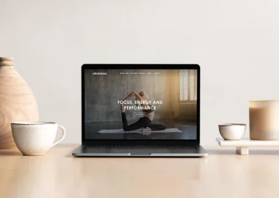 A laptop on a wooden table shows a custom shopify store Australia, with yoga content; vases, candle, plate nearby.