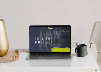A laptop on a white desk shows aresponsive web design Australia. Nearby: black mug, beige cloth, golden vase, white chair.