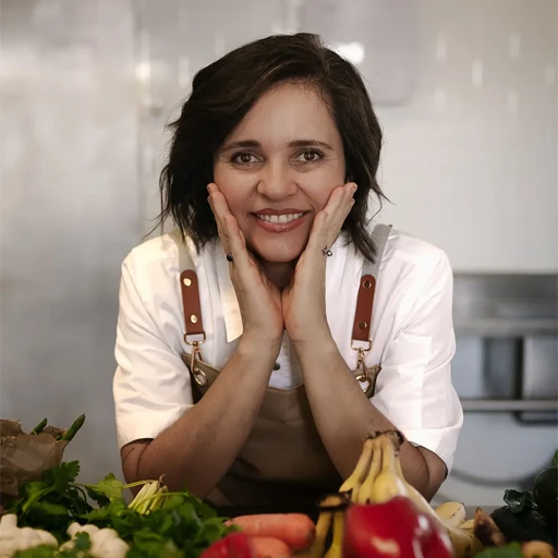 Gabi A woman in a brown apron and white shirt smiles with her hands on her cheeks, leaning forward amid a variety of fresh vegetables on a kitchen counter.