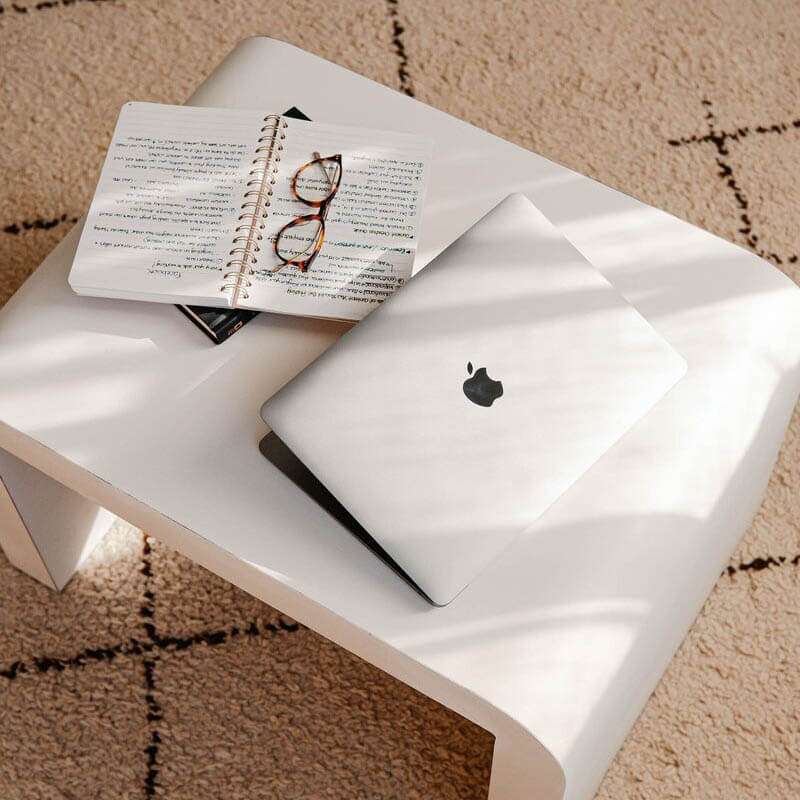 Ingrid-Services-00-1.jpg A closed MacBook and open notebook with glasses on a white table, ideal for online course website design Australia workspace.