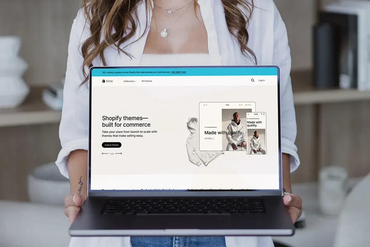 A person holds a laptop showing the Shopify themes webpage with "Shopify themes—built for commerce," featuring Free Shopify Themes.