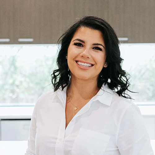 A woman with wavy dark hair, wearing a white button-up shirt and a gold necklace, smiles whilst standing indoors in front of a window and wood-panelled wall. A woman with wavy dark hair, wearing a white button-up shirt and a gold necklace, smiles whilst standing indoors in front of a window and wood-panelled wall.