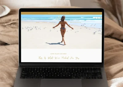 A laptop on a beige bed shows an online shop for a swimwear brand with a woman in a bikini walking on the sandy beach.