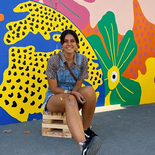 A woman in denim dungarees and a patterned shirt sits on a wooden pallet, smiling in front of a colourful mural with abstract shapes, a green flower, and yellow leopard spots. A woman in denim dungarees and a patterned shirt sits on a wooden pallet, smiling in front of a colourful mural with abstract shapes, a green flower, and yellow leopard spots.
