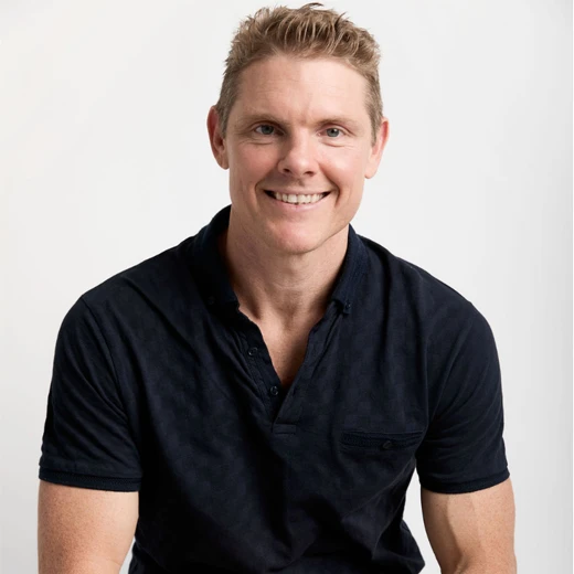 A man with short blond hair, wearing a dark short-sleeved polo shirt, smiles whilst sitting against a plain white background. A man with short blond hair, wearing a dark short-sleeved polo shirt, smiles whilst sitting against a plain white background.