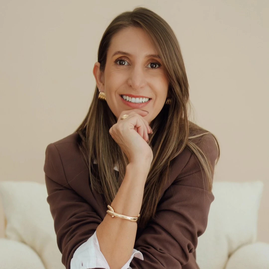 A woman with long brown hair, wearing a brown blazer, white shirt, and gold jewellery, smiles whilst resting her chin on her hand, seated on a light-coloured sofa against a beige background. A woman with long brown hair, wearing a brown blazer, white shirt, and gold jewellery, smiles whilst resting her chin on her hand, seated on a light-coloured sofa against a beige background.