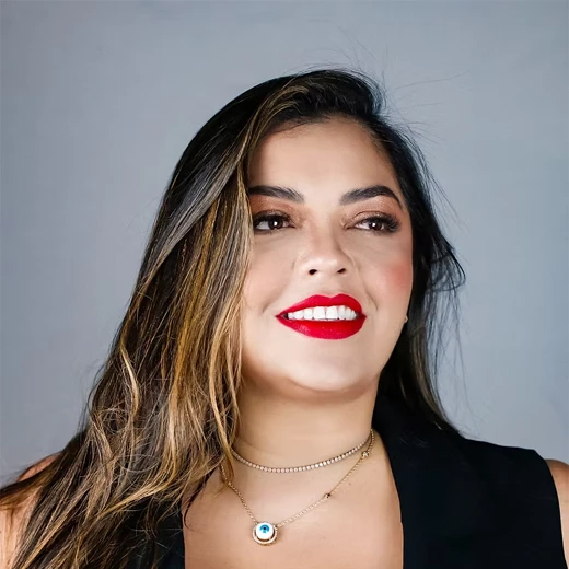 A woman with long brown hair, wearing bright red lipstick, a black sleeveless top, and layered gold necklaces, smiles and looks slightly to the side against a plain grey background. A woman with long brown hair, wearing bright red lipstick, a black sleeveless top, and layered gold necklaces, smiles and looks slightly to the side against a plain grey background.