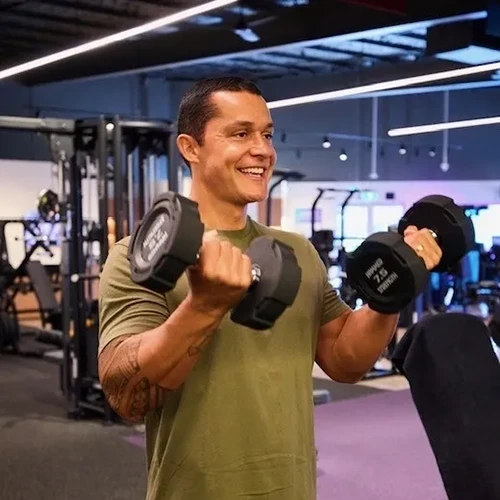 A man in a green shirt smiles whilst lifting two black dumbbells in a gym, surrounded by exercise equipment. A man in a green shirt smiles whilst lifting two black dumbbells in a gym, surrounded by exercise equipment.