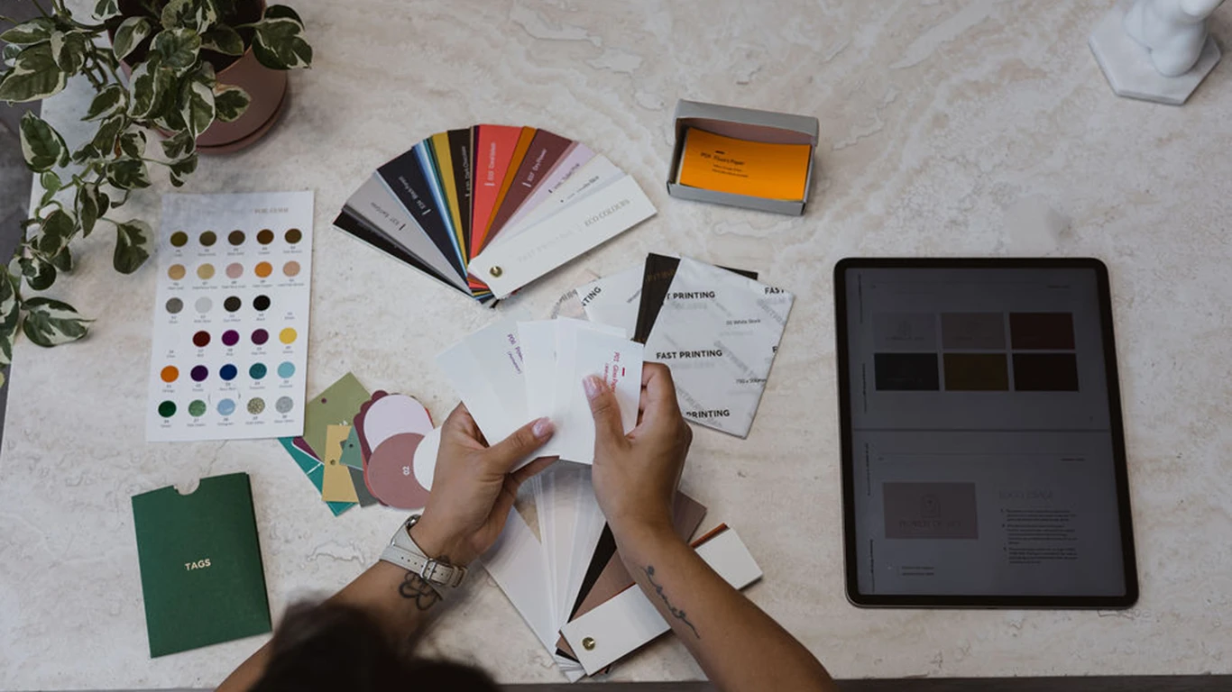 A person reviews colour swatches and printed material samples at a desk with an iPad, sample cards, a plant, and a sheet of colour options. A person reviews colour swatches and printed material samples at a desk with an iPad, sample cards, a plant, and a sheet of colour options.