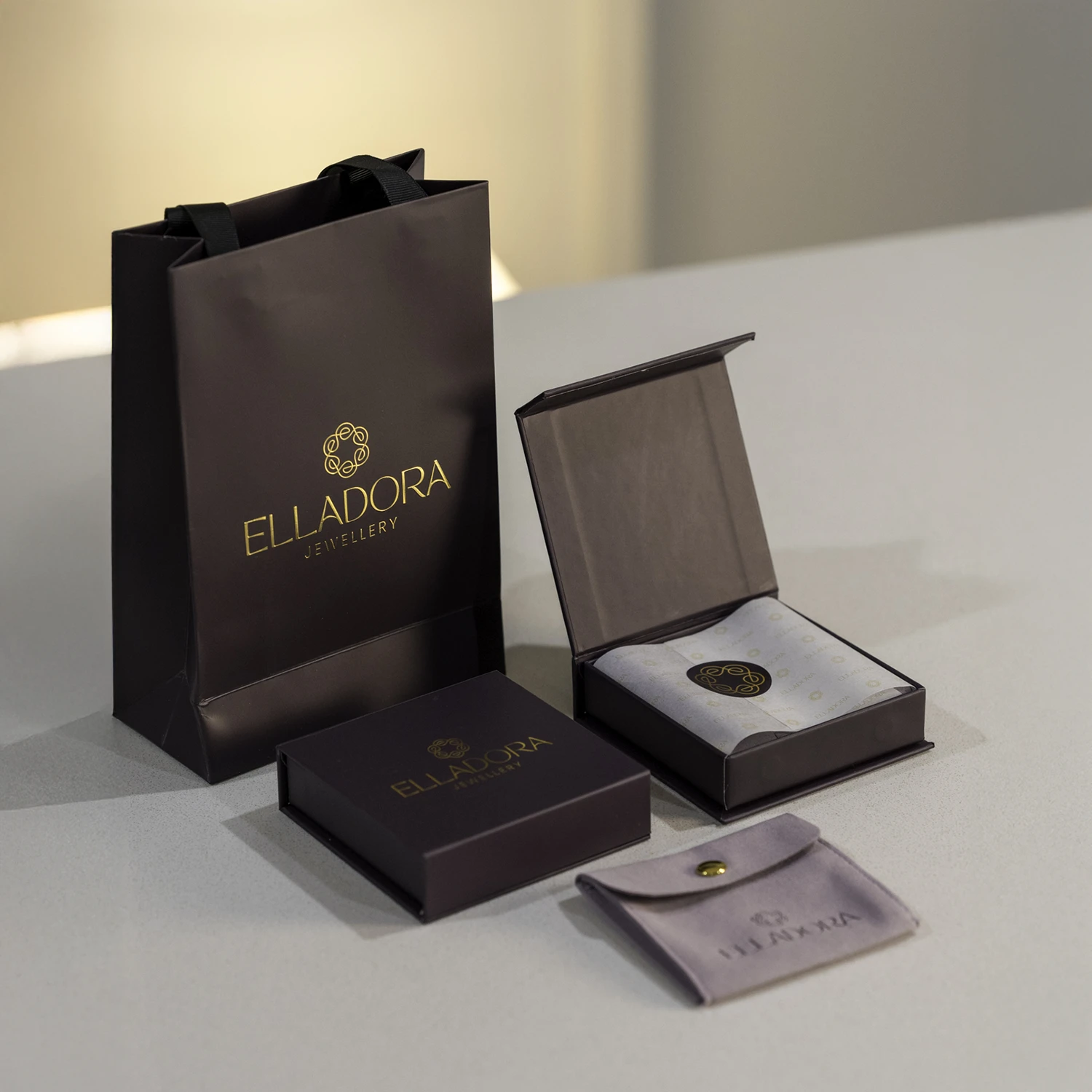 A gift set from Elladora Jewellery featuring a dark purple gift bag, a closed box, an open box with tissue paper, and a small velvet pouch, all with gold Elladora branding, displayed on a light surface. A gift set from Elladora Jewellery featuring a dark purple gift bag, a closed box, an open box with tissue paper, and a small velvet pouch, all with gold Elladora branding, displayed on a light surface.