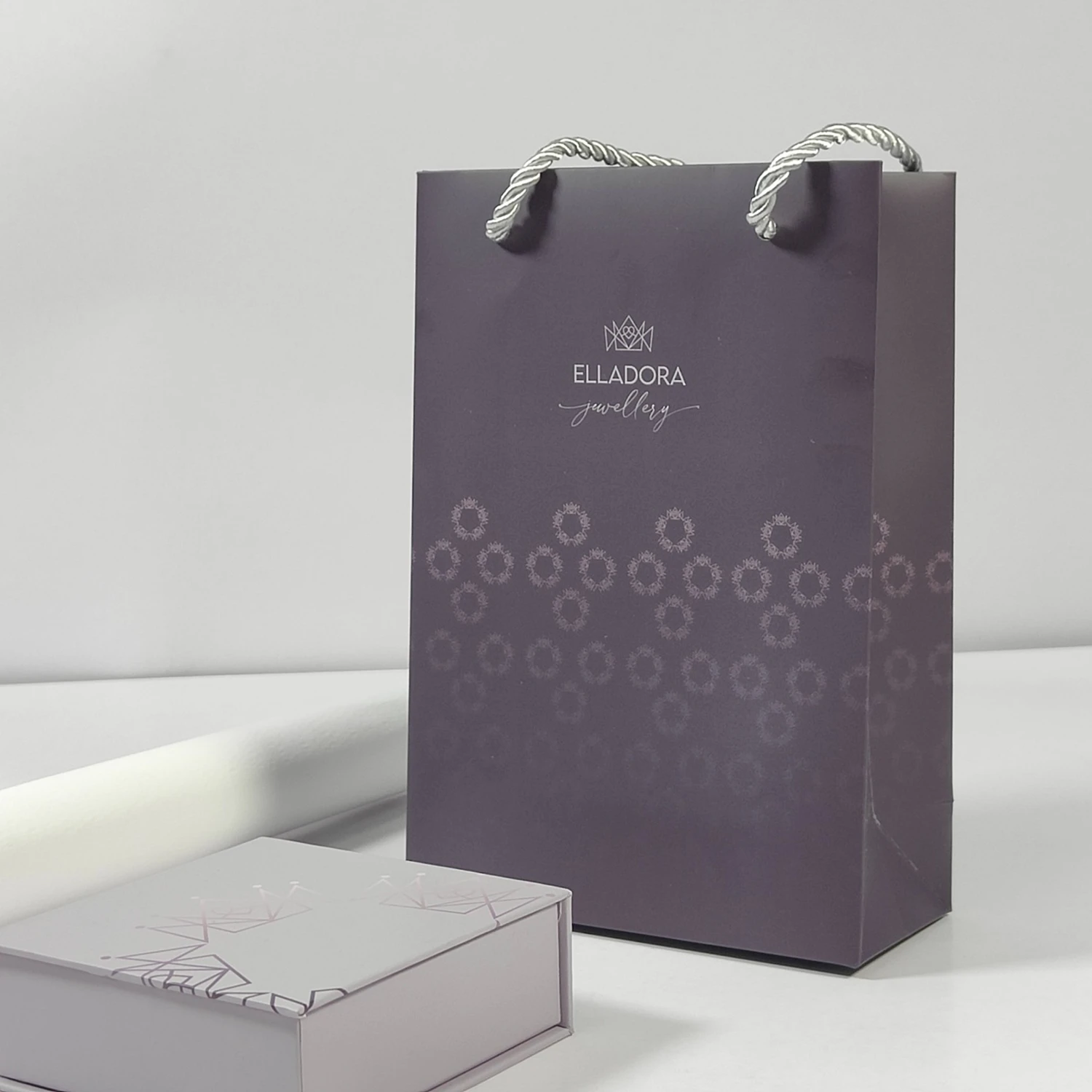 A purple Elladora Jewellery gift bag with white rope handles stands upright next to a matching rectangular box, both featuring subtle geometric patterns and elegant branding, against a plain light background. A purple Elladora Jewellery gift bag with white rope handles stands upright next to a matching rectangular box, both featuring subtle geometric patterns and elegant branding, against a plain light background.